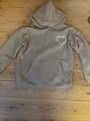 Beige hoodie med hjärtdetalj - Säljer en beige hoodie med huva och magficka. På bröstet finns ett vitt hjärta med ett litet ansikte broderat. Hoodien har ribbade muddar vid ärmar och nederkant. Perfekt för en chill och avslappnad stil.