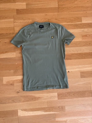 Olivgrön t-shirt från Lyle&Scott - Snygg olivgrön t-shirt med korta ärmar och rund hals. På bröstet sitter en liten broderad logga i gult och svart som ger en clean look. Tillverkad i mjuk bomull för skön känsla och passar perfekt till jeans eller shorts.
