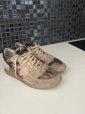Beige sneakers med camomönster - Snygga sneakers i beige mocka och textil med camouflagemönster i brunt och grönt. Skorna har låga skaft, beige snörning och en klassisk gummisula. Perfekta för dig som vill ha en unik och trendig look.
