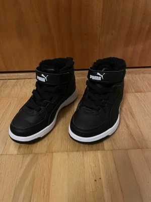 Svarta höga sneakers från Puma - Snygga svarta höga sneakers från Puma med vit sula och svart fuskpälsfoder. Skorna har kardborreband upptill och klassiska svarta skosnören. Perfekta för dig som vill ha både stil och komfort under kalla dagar.