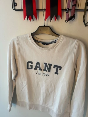 Vit sweatshirt från GANT - Klassisk vit sweatshirt från GANT med tryckt logga och texten 'Est. 1949' på bröstet. Tröjan har rund halsringning, långa ärmar och ribbade muddar. Perfekt för en avslappnad och stilren look.
