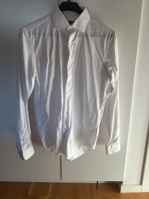 Vit skjorta från Dressmann Slim Fit - Klassisk vit skjorta från Dressmann i slim fit-modell. Tillverkad i stretchig bomull för extra komfort. Skjortan har långa ärmar, knappar framtill och en stilren krage. Perfekt för dig som gillar en clean och enkel look.
