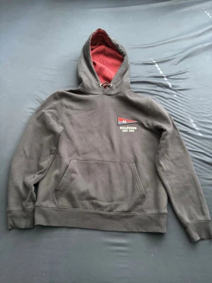 Tommy hilfiger hoddie svart - Säljer en svart hoodie med röd insida i huvan och en liten logga med texten 'HILFIGER EST. 1985' på bröstet. Hoodien har känguruficka framtill och ribbade muddar vid ärmar och nederkant. Perfekt för en chill och avslappnad stil.