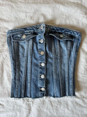 Blå jeanskorsett med knappar - Trendig blå jeanskorsett med silverfärgade knappar framtill och rå kant nertill. Toppen har två dekorativa bröstfickor och smockad rygg för extra komfort. Perfekt för dig som vill ha en edgy och unik look.