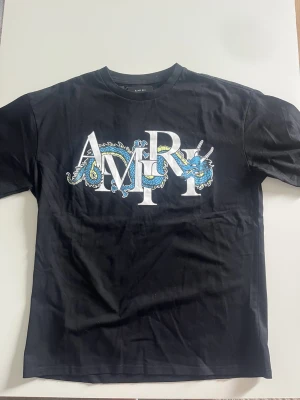 Svart AMIRI t-shirt - Säljer en svart t-shirt från AMIRI med stort tryck framtill där AMIRI-loggan är dekorerad med en blå och gul drake. T-shirten har rund hals och korta ärmar. Tillverkad i mjuk bomull för en skön känsla och avslappnad passform.