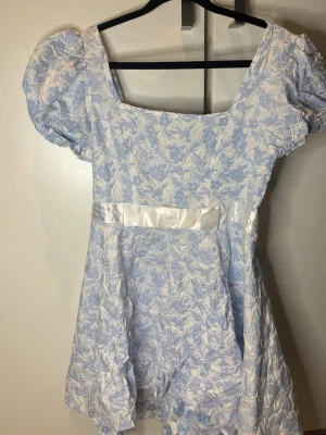 Blåvit blommig kortklänning - Söt blåvit kortklänning från Shein med puffärmar och blommigt mönster. Klänningen har en markerad midja med ett vitt satinband och fyrkantig urringning. Perfekt för sommarens alla tillfällen! Använd endast en gång! Behöver strykas men utöver det jättefin🙂