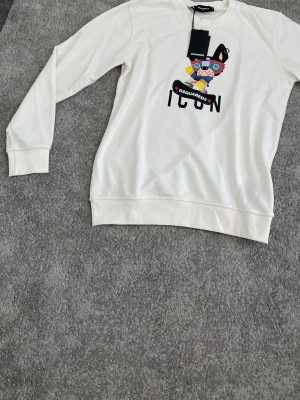 Vit sweatshirt Dsquared2 ICON - Säljer en vit sweatshirt från Dsquared2 med färgglad tryckt grafik och texten ICON på bröstet. Tröjan har rund hals, långa ärmar och ribbade muddar vid ärmslut och nederkant. Perfekt för dig som gillar streetwear och vill sticka ut.
