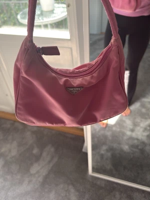 Rosa Prada väska - Trendig rosa handväska från Prada i nylon med klassisk axelrem och ikonisk metallogo framtill. Väskan har en mjuk, rundad form och stängs med dragkedja. Perfekt accessoar för att lyfta din outfit med en poppig färg.
