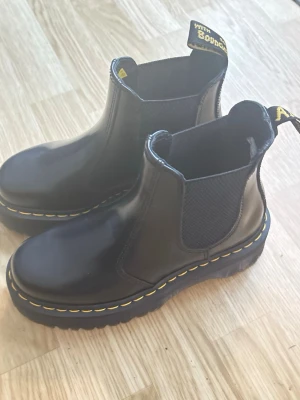 Svarta Chelsea boots med gul söm - Säljer ett par svarta Chelsea boots från Dr. Martens med klassisk gul söm runt sulan och elastiska paneler på sidorna. Skorna har en rejäl platåsula och dragflikar med gul text bak. Perfekta för dig som vill ha en edgy och tidlös look.