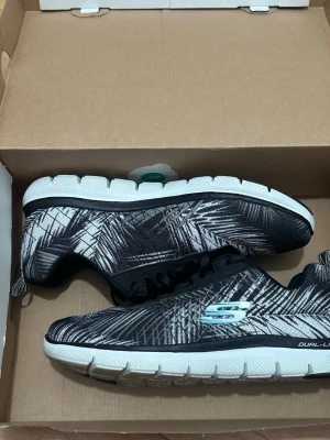 Svarta Skechers sneakers med palmtryck - Snygga svarta Skechers sneakers med vitt palmbladsmönster och vit sula. Skorna har en lätt och flexibel design med svart snörning och Skechers-logga i ljusblått på sidan. Perfekta för dig som vill ha både stil och komfort i vardagen.