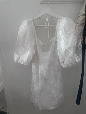 Vit kort spetsklänning med puffärm - Superfin vit kort klänning med puffiga ärmar och vacker spets med blommigt mönster. Klänningen har öppen rygg med knytband i nacken och romantisk känsla. Perfekt för sommarens alla tillfällen. Har enbart använt klänningen på min student! 