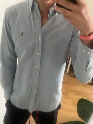Ljusblå skjorta från Ralph Lauren - Klassisk ljusblå skjorta från Ralph Lauren med broderad logga på bröstet. Skjortan har knappar framtill, button-down krage och lång ärm. Tillverkad i mjuk bomull och har en normal passform som ger en clean och stilren look.