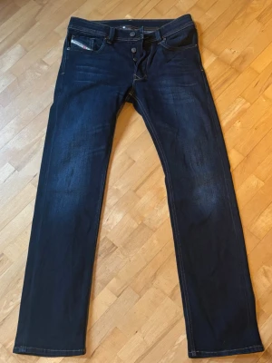Mörkblå raka jeans med knappar - Säljer ett par mörkblå jeans med raka ben och knappgylf. Jeansen har klassiska fem fickor och kontrastsömmar. Materialet är denim och passformen är normal. Perfekta för dig som gillar en enkel och stilren look.