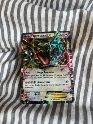 Rayquaza EX Pokémonkort - Säljer ett Rayquaza EX Pokémonkort med holografisk yta. Kortet har färgstark design med Rayquaza i svart, grönt och gult samt detaljer i rosa och blått. Kortet har 170 HP och attacker som Mega Ascension och Aeroscream. Perfekt för samlare eller Pokémonfans.
