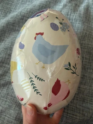 Stort påskägg i papp med hönsmotiv - Säljer ett stort påskägg i papp med färgglada illustrationer av hönor, kycklingar och blommor. Ägget har en ljus bottenfärg och detaljer i blått, gult, rött och lila. Perfekt för att fylla med godis eller små presenter till påsk.