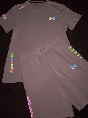 Under Armour grått träningsset - Säljer ett grått träningsset från Under Armour med t-shirt och shorts. Setet har färgglada loggor och detaljer i regnbågsfärger, samt elastisk midja med dragsko på shortsen. Perfekt för gymmet eller löpning.