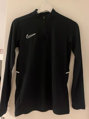 Svart Nike träningsjacka med dragkedja - Snygg svart träningsjacka från Nike med vit logga på bröstet och halv dragkedja framtill. Jackan har lång ärm och vita detaljer vid sidorna. Perfekt för träning eller uppvärmning. Tillverkad i ett stretchigt funktionsmaterial.
