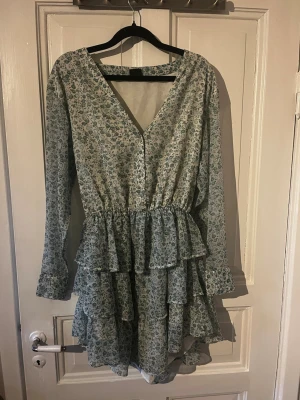 Blommig volangklänning Gina Tricot XS - Supersöt kort blommig klänning från Gina Tricot i ljusblått och vitt. Klänningen har v-ringning, långa ärmar och flera volanglager i kjolen. Perfekt för sommarens alla tillfällen och ger en romantisk vibe.