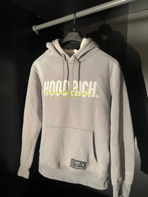 Grå hoodie från Hoodrich - Snygg grå hoodie från Hoodrich med stor vit och gul broderad logga på bröstet. Klassisk känguruficka framtill, justerbar huva med dragsko och ribbade muddar. Perfekt för en avslappnad streetwear-look.
