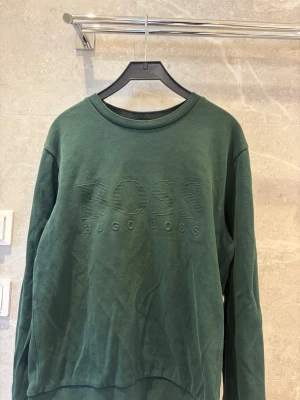Grön sweatshirt från Hugo Boss - Mörkgrön sweatshirt från Hugo Boss med broderad logga på bröstet. Tröjan har rund halsringning, långa ärmar och ribbade muddar. Slim fit-modell som ger en snygg och modern siluett. Perfekt för en avslappnad och stilren look.Storlek M men S kan också ha den.