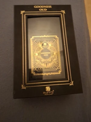 Goodness Oud parfym från RiiFFS - Lyxig parfymflaska från RiiFFS med namnet Goodness Oud. Flaskan är svart med detaljer i guld och har en elegant, rektangulär form. Förpackningen är svart med guldfärgade mönster och text, vilket ger en exklusiv känsla. Perfekt för dig som gillar orientaliska dofter.