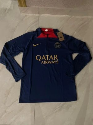 Mörkblå PSG träningströja Nike - Snygg långärmad träningströja från Paris Saint-Germain i mörkblått med guldfärgade detaljer och logga. Halvlång dragkedja framtill och röd insida vid kragen. Tillverkad i ett lätt och funktionellt material, perfekt för fotbollsträning.