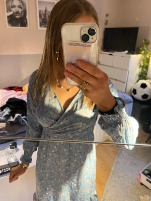 Blommig ljusblå omlottklänning - Söt ljusblå omlottklänning med små blommor i vitt och rosa. Klänningen har långärmade ärmar med smock vid handlederna och v-ringad hals. Perfekt för vår och sommar, ger en romantisk och fräsch look.