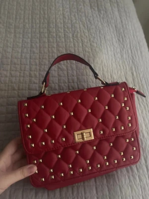 Röd quiltad handväska med nitar - Snygg röd handväska i quiltad design med guldfärgade nitar och vridlås. Väskan har ett kort handtag upptill och detaljer i guld. Perfekt accessoar för att lyfta din outfit.