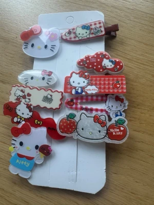 Hello Kitty hårspännen mix - Säljer ett set med färgglada hårspännen med Hello Kitty-motiv. Spännena har olika former och mönster, bland annat rutor, äpplen och blommor i rött, vitt och rosa. Perfekt för dig som gillar söta accessoarer och vill piffa till frisyren med lite extra kawaii.