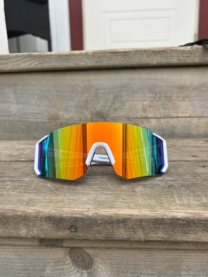 Orange Vita sportiga solglasögon med spegelglas - Säljer ett par riktigt feta solglasögon med vit båge och stora spegelglas i regnbågsfärger. Glasen har en futuristisk, kantig form och ger en cool vibe till alla outfits. Perfekta för dig som vill sticka ut och gillar sportig stil.