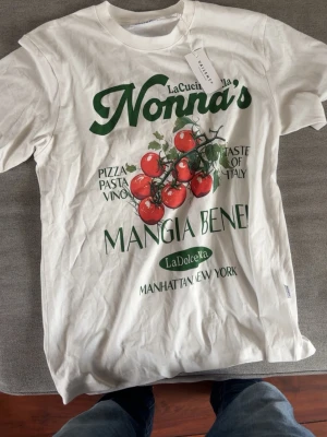 Vit t-shirt med tomattryck från Vailent - Snygg vit t-shirt från Vailent med stort grönt och rött tryck framtill. Motivet har tomater på kvist och italiensk text som ger en retrovibe. Rund halsringning och korta ärmar. Perfekt för dig som gillar streetwear och grafiska prints.