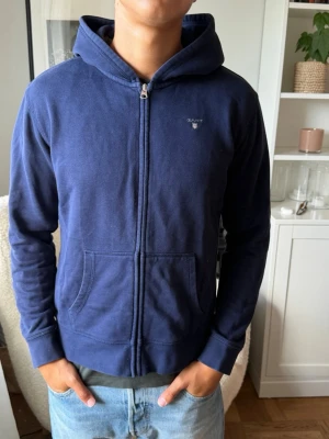Mörkblå zip hoodie från Gant - Snygg mörkblå hoodie från Gant med dragkedja framtill och två fickor. Klassisk huva och diskret Gant-logga på bröstet. Tillverkad i mjukt bomullsmaterial som känns skönt mot huden. Perfekt för en avslappnad och stilren look. L i barn