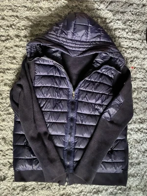 Mörkblå Moncler cardigan med huva - Säljer en mörkblå Moncler cardigan pufferjacka med quiltad front och rygg i glansigt material samt stickade ärmar och muddar. Jackan har dragkedja framtill och en stor huva som ger extra skydd mot kyla. Perfekt för dig som vill ha en stilren och varm jacka till vintern.
