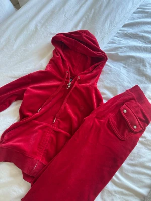 Juicy Couture, rött velourset - Mysigt loungewear-set i röd velour med matchande hoodie och byxor. Hoodien har dragkedja, fickor och snörning i huvan. Byxorna har resår i midjan och ficka med lock och knappar. Perfekt för chill dagar hemma.