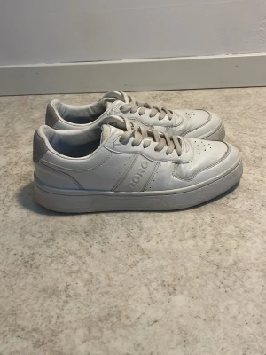 Vita sneakers från Björn Borg - Snygga vita sneakers från Björn Borg med grå detaljer på hälen och perforeringar på tån. Skorna har en klassisk låg siluett, snörning och är tillverkade i läderliknande material. Perfekta för en clean och stilren look.