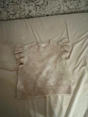 Beige stickad väst med volangärmar - Supersöt beige stickad väst med volangdetaljer vid axlarna och ribbstickade kanter. Perfekt att styla över en skjorta eller t-shirt för en trendig lager-på-lager-look. Mjuk och skön, passar till många outfits.