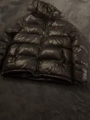 Svart dunjacka från Polo Ralph Lauren - Snygg svart dunjacka från Polo Ralph Lauren med glansig finish och röd broderad logga på bröstet. Jackan har huva, dragkedja framtill och två sidofickor med dragkedja. Perfekt för kalla vinterdagar. Passar mellan 170-185 riktigt skön och inga diirekta skavanker på den mer än använd i ett i ett halvår bara höra av sig vid frågor🙃