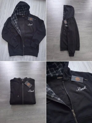 Svart hoodie med rutigt foder och dragkedja - Säljer en svart hoodie med dragkedja och huva. Insidan av huvan har ett rutigt mönster i grått och svart. På bröstet finns en vit broderad text 'Marcel'. Hoodien har dragsko i huvan och ribbade muddar vid ärmar och nederkant.