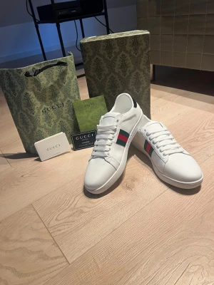 Vita Gucci Ace sneakers med bi - Snygga vita Gucci Ace sneakers i läder med klassiska gröna och röda ränder på sidan samt en guldbroderad bi. Skorna har vita snören och låg profil, perfekt för dig som vill ha en clean och lyxig look. Kommer med originalförpackning och tillbehör.