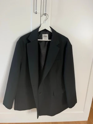 Svart kavaj från Zara - Stilren svart kavaj från Zara med klassisk passform och enkelknäppning. Kavajen har breda slag, två framfickor och en bröstficka. Tillverkad i ett slätt tyg med helfodrad insida