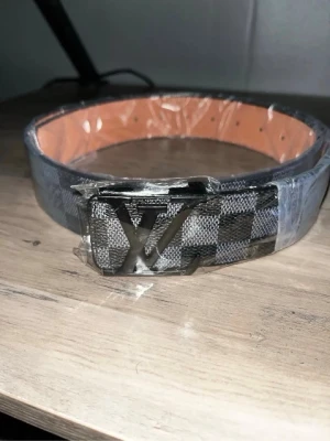 Rutigt Louis Vuitton bälte i skinn - Säljer ett Louis Vuitton bälte med klassiskt rutigt mönster i svart och grått. Bältet har en stor LV-logga i metall som spänne och insidan är i ljust skinn. Perfekt accessoar för att lyfta din outfit.