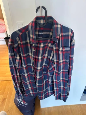 Rutig skjorta från Jaktia, medium - Säljer en klassisk rutig skjorta från Jaktia i storlek medium. Skjortan har blå, röda och vita rutor, lång ärm och knappar framtill. Perfekt för dig som gillar en avslappnad men ändå stilren look.