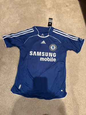 Chelsea Retro tröja #11 Drogba - Blå Chelsea matchtröja från Adidas med vita detaljer och tryck. Framsidan har Samsung Mobile-logga och Chelsea FC-märke, baksidan har DROGBA och nummer 11. Klassisk fotbollströja i funktionsmaterial, perfekt för fans och samlare.