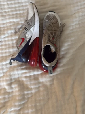 Nike Air Max 270 beige/röd sneakers - Snygga Nike Air Max 270 sneakers i beige mesh med röda och svarta detaljer samt en stor röd luftbubbla i sulan. Skorna har en sportig look med ventilerande material och klassisk Nike Swoosh på sidan. Perfekta för dig som gillar streetwear och komfort.