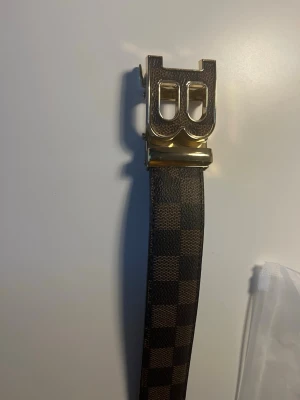 Rutigt brunt BB bälte - Snyggt bälte med rutigt mönster i bruna och svarta toner. Bältet har ett stort, guldigt B-format spänne som ger en lyxig känsla. Perfekt accessoar för att lyfta din outfit och ge den en trendig touch.