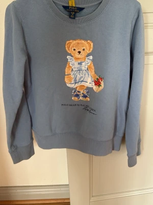Blå stickad tröja Polo Ralph Lauren - Supersöt blå stickad tröja från Polo Ralph Lauren med ikoniska Polo Bear-motivet framtill. 