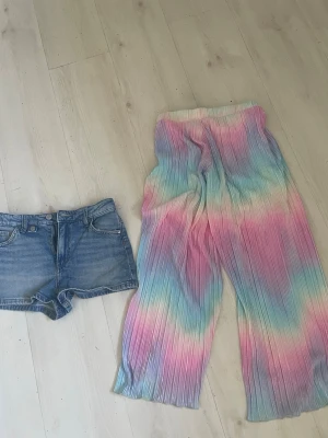 Blå jeansshorts med klassisk look+Regnbågs byxor - Säljer ett par blå jeansshorts med fem fickor och knappgylf. Shortsen har en klassisk denimlook och är perfekta för varma dagar. Passar dig som gillar en enkel och tidlös still. Regnbågsbyxor inutsvängd nedbhlittet. Fodrade 