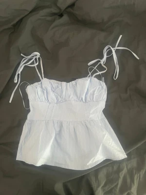 Ljusblå topp med knytband från Gina Tricot - Supersöt ljusblå topp från Gina Tricot med tunna vita ränder och knytbara axelband. Toppen har en rynkad byst och smockad rygg för skön passform. Tillverkad i ett lätt bomullsliknande material, perfekt för sommardagar.