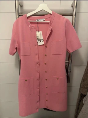 Rosa kort klänning från Zara - Säljer en söt rosa kort klänning från Zara med korta ärmar, rund halsringning och dekorativa guldknappar framtill. Klänningen har fyra fickor på framsidan och är gjord i ett mjukt, stickat material. Perfekt för dig som gillar en chic och enkel stil.