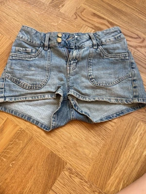Blå denimshorts med broderade fickor - Säljer ett par blå denimshorts med två framfickor och dubbla knappar i midjan. Baksidan har två fickor med snyggt broderade hjärtan. Klassisk jeanslook med ljus tvätt och bälteshällor. Perfekta för varma dagar!